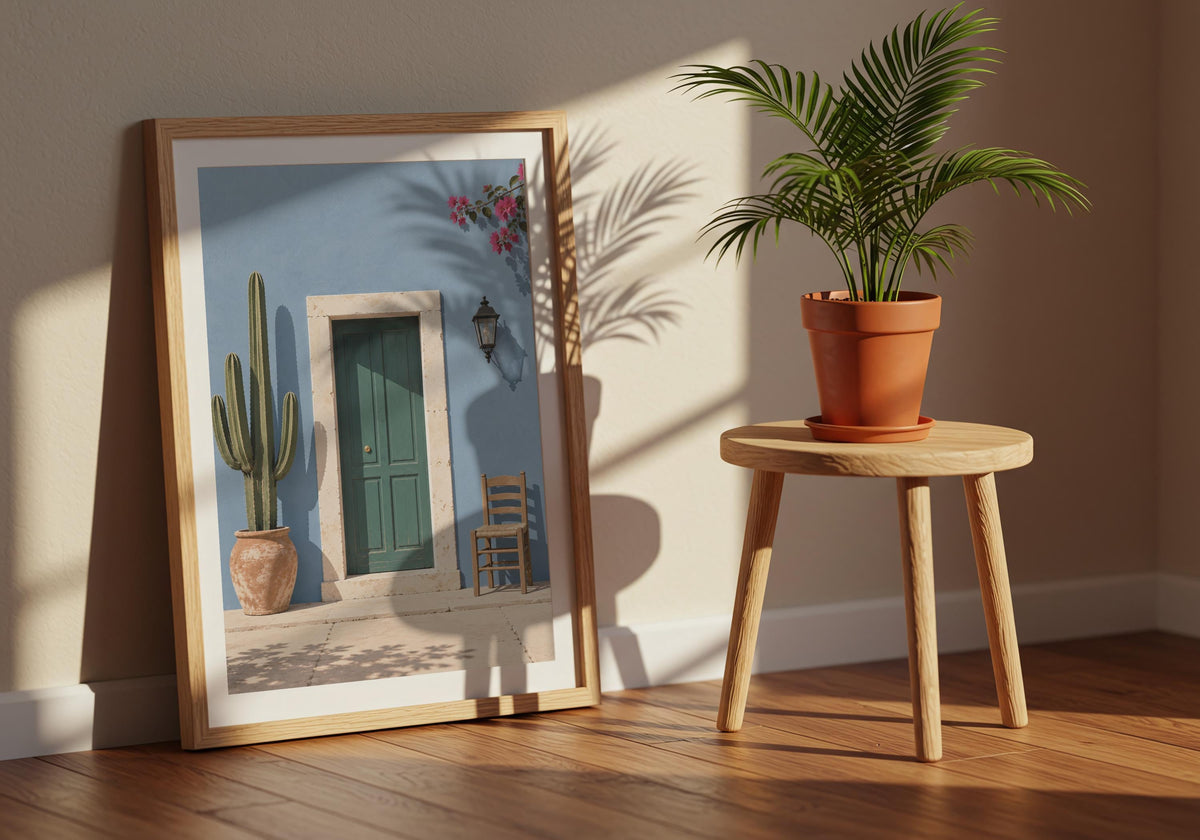 Angled view of entryway wall art depicting a forest-green door in a powder-blue wall with cactus, chair and flowers, leaning in a dark wood frame beside a terracotta plant.