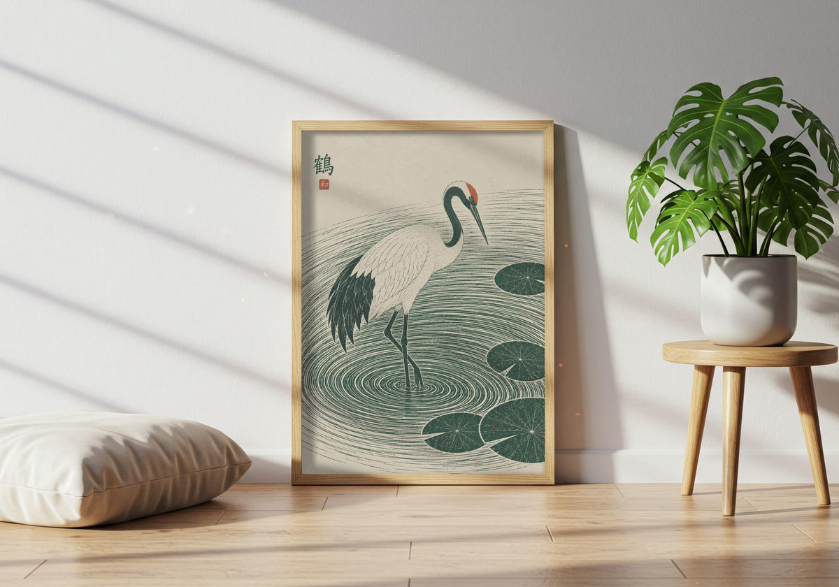 Japanese wall art featuring a white crane stepping through deep green ripples and lily pads, leaning in a light wood frame near a healthy green plant.