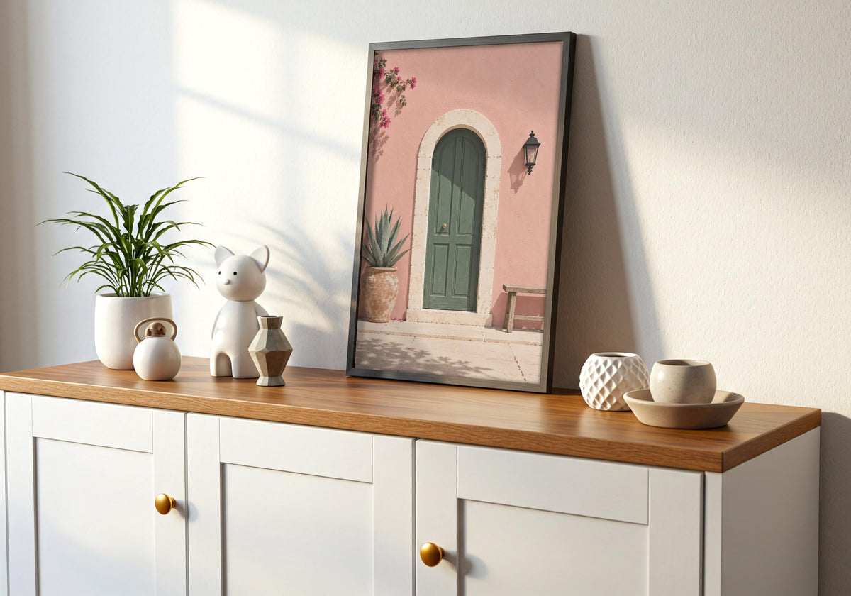 Interior mockup of mediterranean art depicting an arched green door in a blush-pink wall with bougainvillea and a wooden bench, displayed in a black frame on a wooden console.