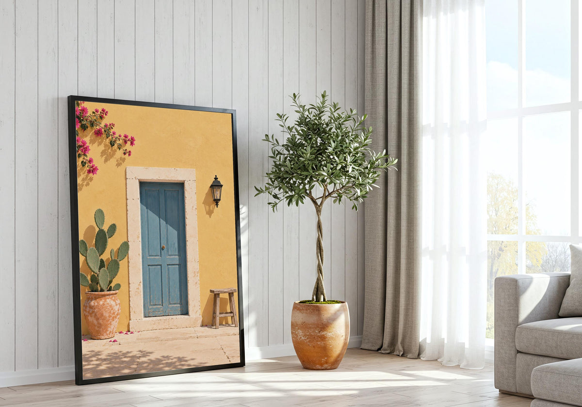 Yellow Wall Blue Door presented as yellow wall art, featuring a dusty-blue door in a sunny yellow wall with cactus, stool and bougainvillea and hung in a black frame beside a bright window.