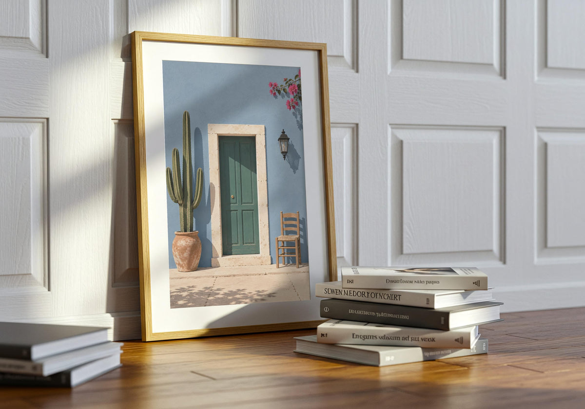 Colourful home art featuring a forest-green door in a powder-blue wall with cactus, chair and flowers, styled in a light wood frame on stacked books.