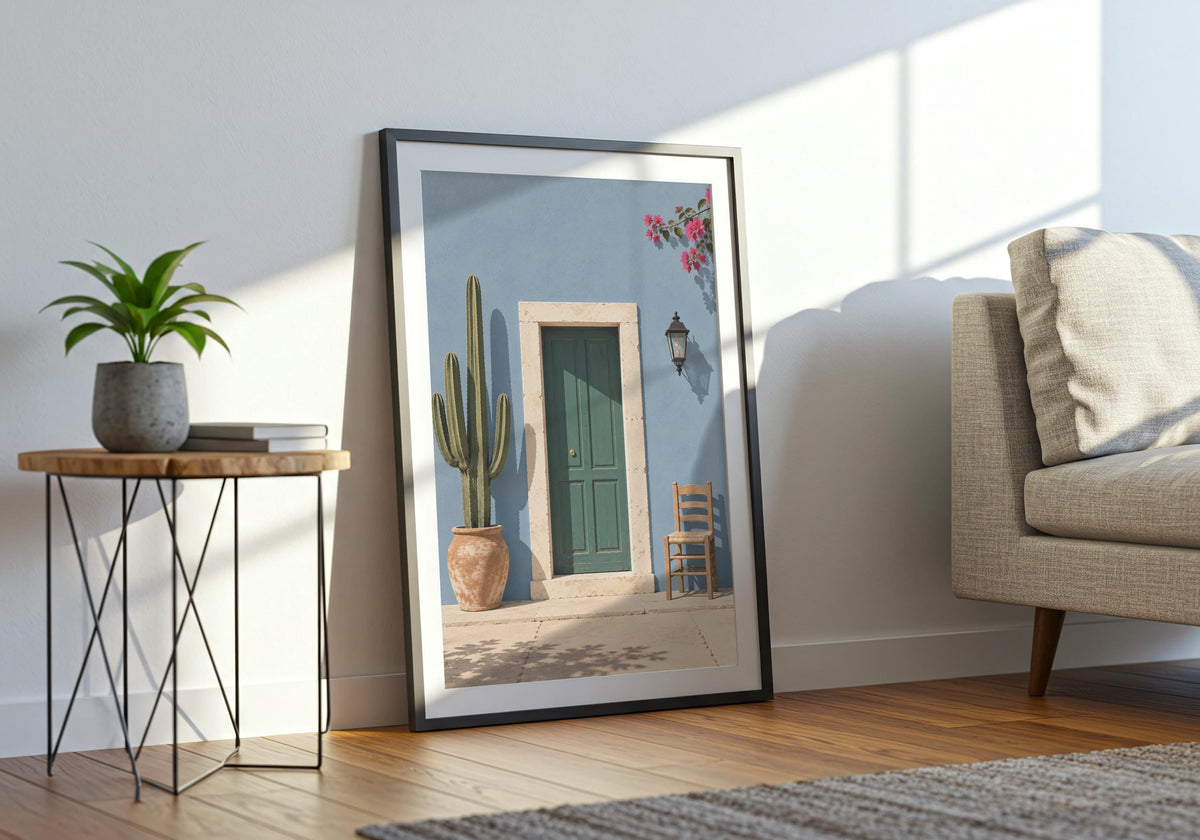 Wide interior view of summer wall decor with a forest-green door in a powder-blue wall with cactus, chair and flowers, shown in a black frame beside a pale sofa.