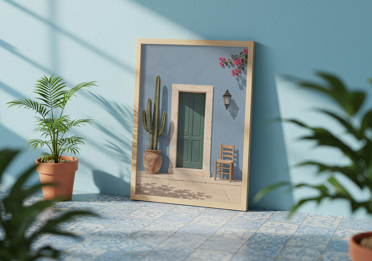 Travel wall decor showing a forest-green door in a powder-blue wall with cactus, chair and flowers, displayed in a pale frame among leafy plants.