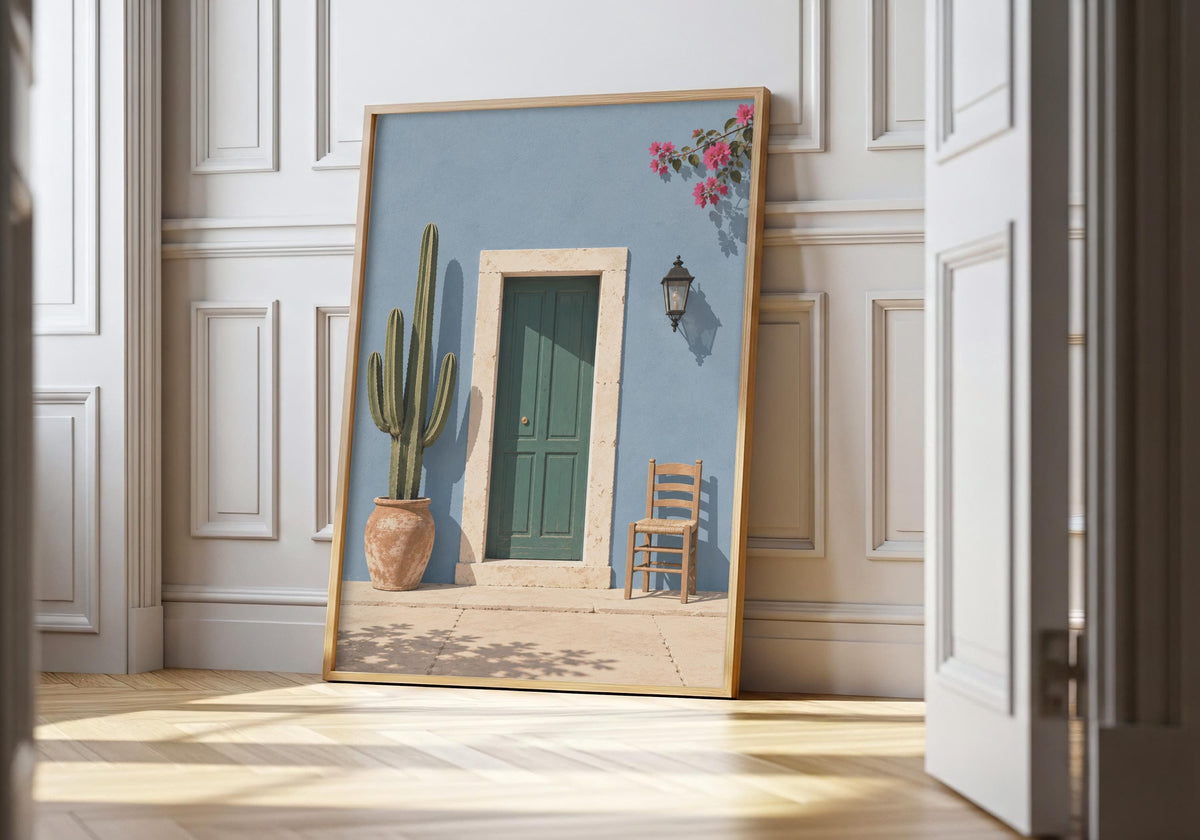 Green door print showing a forest-green door in a powder-blue wall with cactus, chair and flowers, leaning in a light natural-wood hero frame against white panelling.
