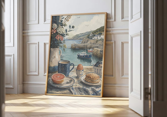 Cornish wall art showing coffee, toast and an egg overlooking boats in a calm Cornish harbour, leaning in a light natural-wood hero frame against white panelling.