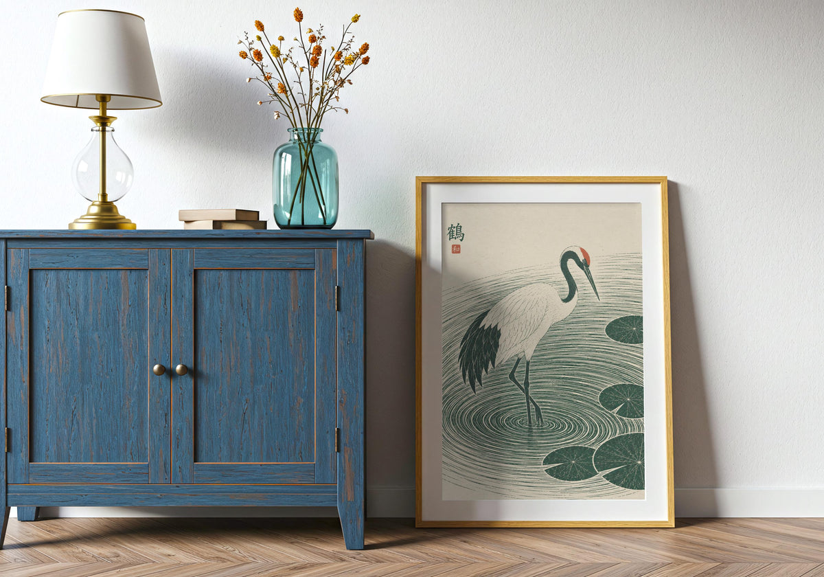 Japandi wall art with a white crane stepping through deep green ripples and lily pads, leaning in a light wood frame beside a blue cabinet.