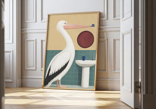 Pelican wall art showing a white pelican carrying a toothbrush beside a tiled sink and round mirror, leaning in a light natural-wood hero frame against white panelling.