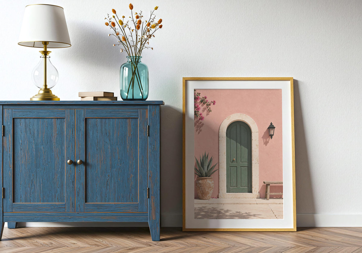 Doorway wall print with an arched green door in a blush-pink wall with bougainvillea and a wooden bench, leaning in a light wood frame beside a blue cabinet.