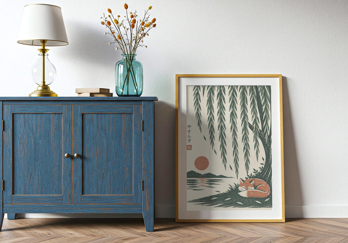 Japanese style art with a rust-coloured fox curled asleep beneath cascading willow branches and a low sun, leaning in a light wood frame beside a blue cabinet.