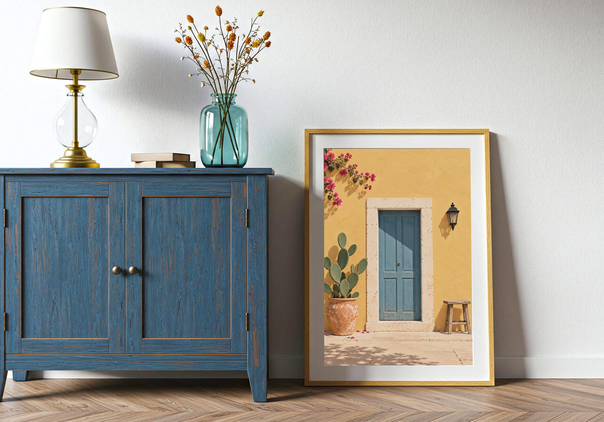 Doorway wall print with a dusty-blue door in a sunny yellow wall with cactus, stool and bougainvillea, leaning in a light wood frame beside a blue cabinet.