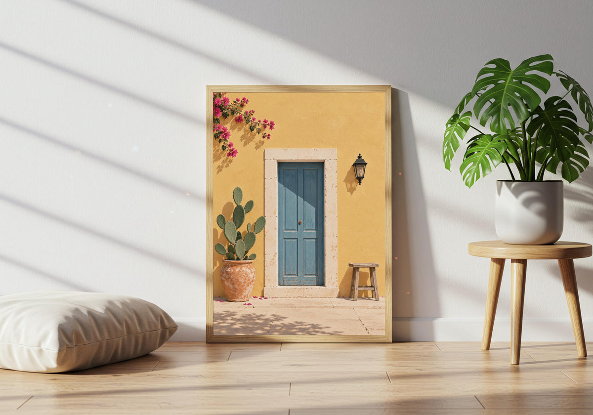 Colourful home art featuring a dusty-blue door in a sunny yellow wall with cactus, stool and bougainvillea, leaning in a light wood frame near a healthy green plant.