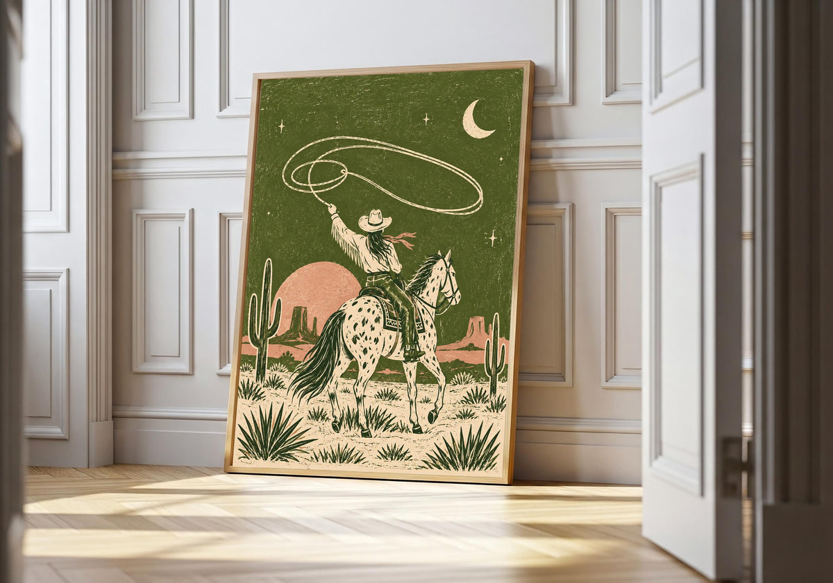 Desert Cowboy Lasso cowboy wall art in a light wood frame, featuring a spotted-horse cowboy twirling a lasso beneath a crescent moon in an olive desert.