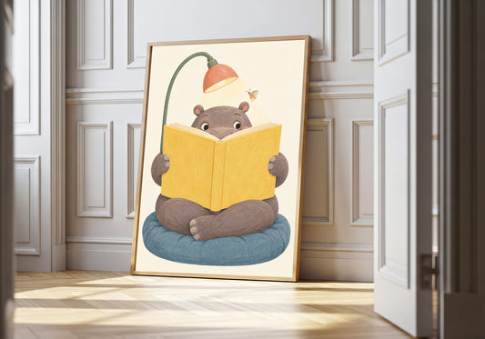 Hippo's Quiet Hour hippo nursery art in a light wood frame, featuring a gentle hippo seated on blue cushions reading a large yellow book beneath a curved lamp.