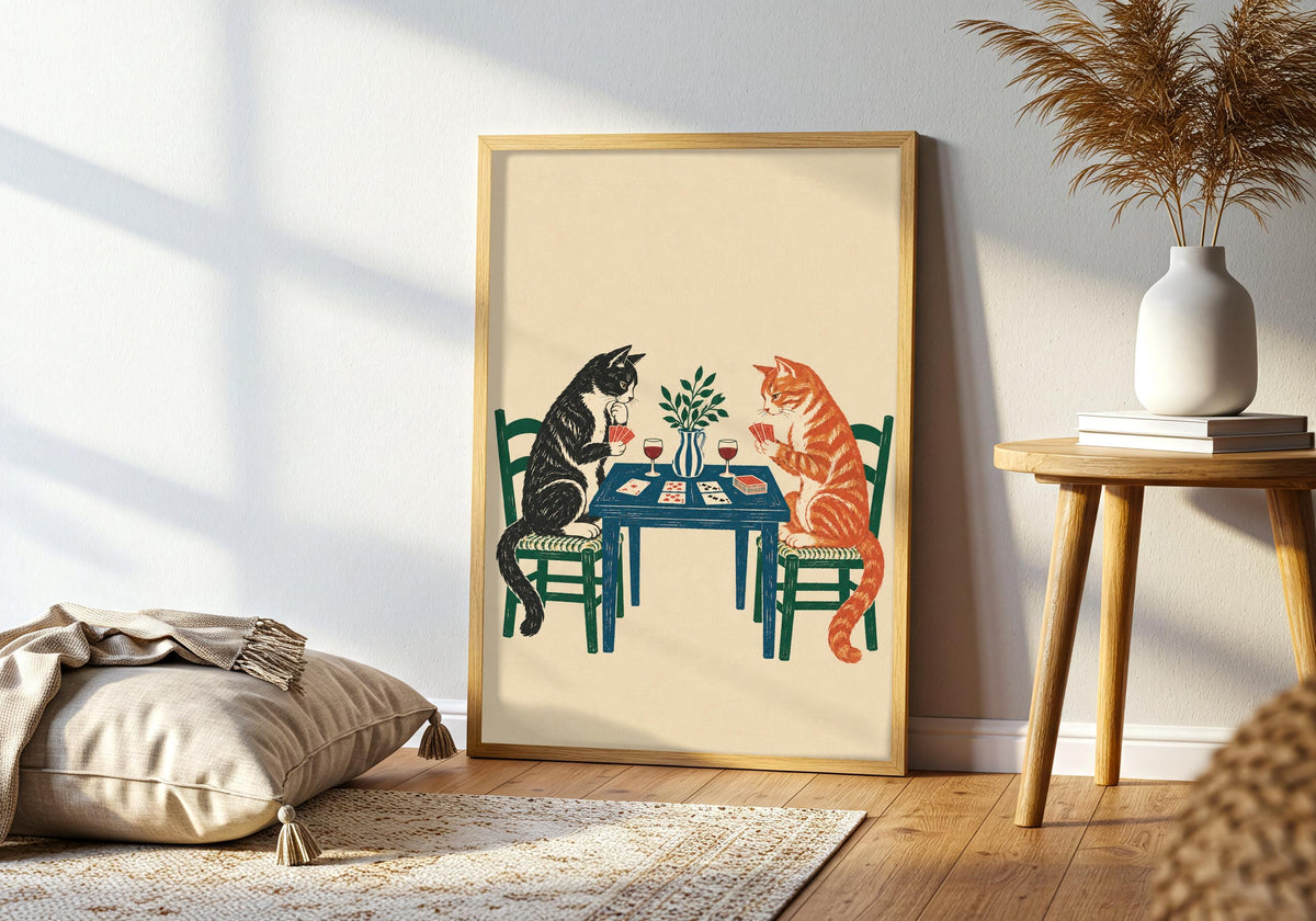 Greenery and furniture surround the framed artwork depicting a tuxedo cat and ginger tabby playing cards over wine at a blue table.