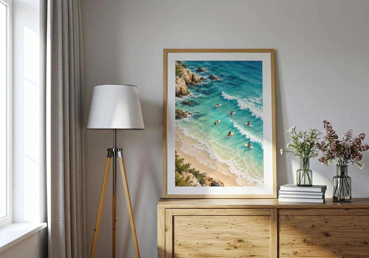 Placed above a wooden chest beside a tripod lamp, the artwork shows small surfers in turquoise water.